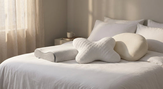 Three distinctly shaped cervical pillows — butterfly, contour, and shredded foam — arranged on white bedding in a softly lit, serene bedroom setting.