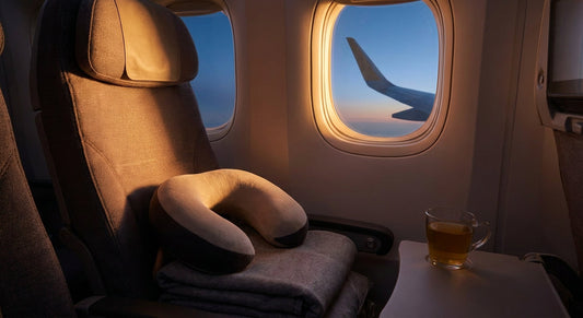 A memory foam travel pillow resting on an airplane window seat bathed in warm golden light, suggesting comfort and cervical spine support during long-haul travel.